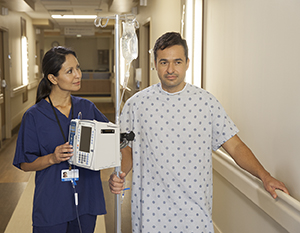 Paciente que camina por el pasillo de un hospital con un soporte intravenoso junto a una proveedora de atención médica.