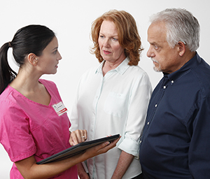 Proveedora de atención médica sosteniendo una tableta electrónica mientras habla con una mujer y un hombre.